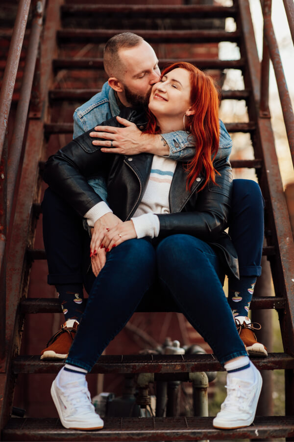 An intimate moment between a couple seated on an industrial staircase, with the man embracing the woman from behind, set in an urban, rustic environment.