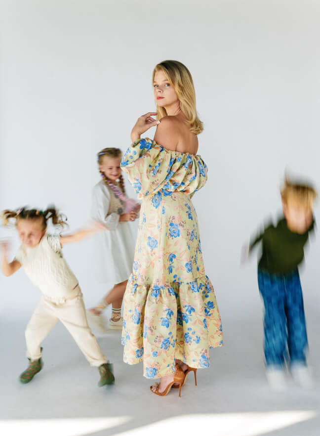 A woman in a flowery, off-the-shoulder dress standing poised with children in motion around her.