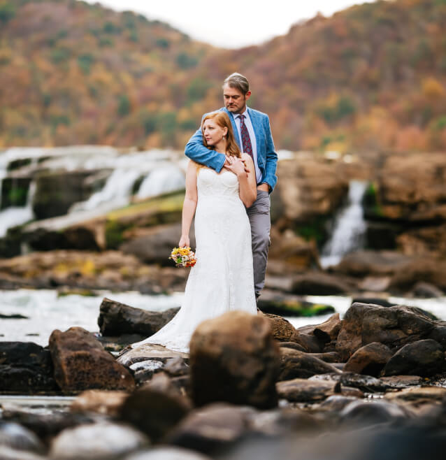 A bride and groom standing amidst a rugged landscape of rocks and a waterfall backdrop, with the bride in a classic white dress and blue shawl, and the groom in a gray suit with a blue tie.