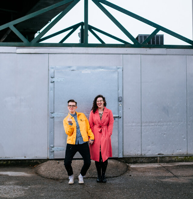 Two people in bright outerwear, a yellow jacket and a pink coat, stand smiling before a metallic industrial door, with a geometric green steel structure overhead.