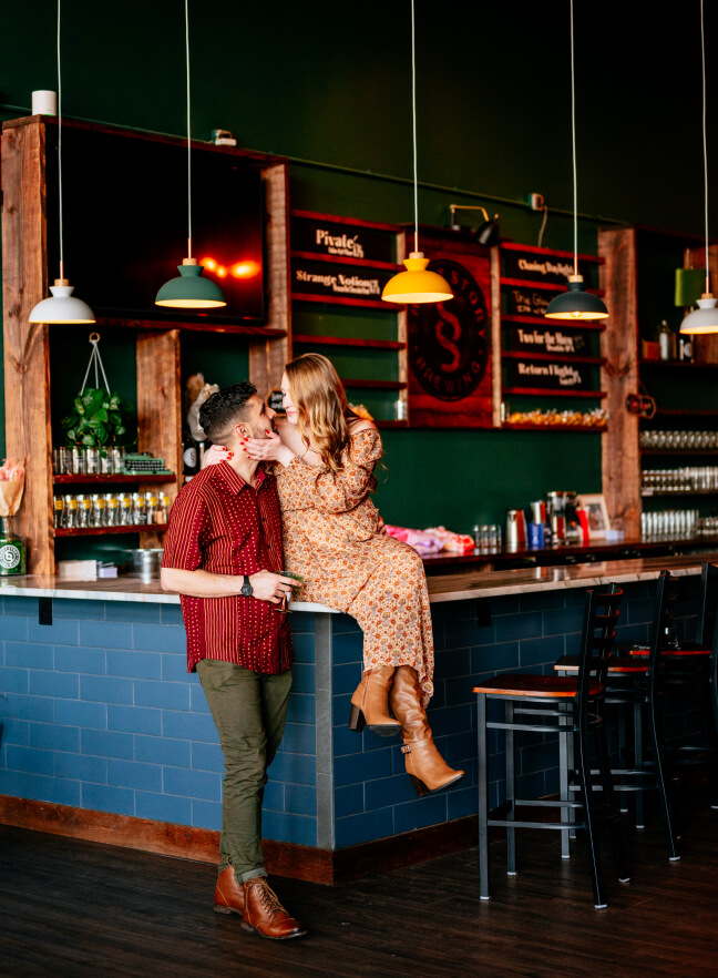 A couple sharing a romantic moment at a cozy bar, with the woman sitting on the counter in a floral dress and the man in a burgundy shirt, surrounded by warm lighting and rustic decor.