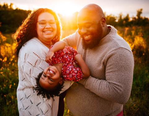 A joyful family moment captured at golden hour, with a beaming couple swinging their delighted child between them, the warmth of the sunset enhancing their smiles.