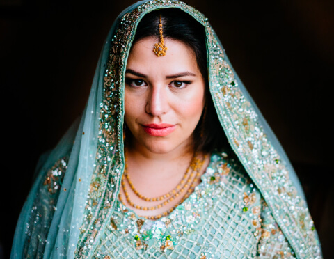 A portrait of a woman adorned in a traditional South Asian bridal attire with intricate embroidery, her expression serene and contemplative, against a dark background that accentuates the sparkle of her outfit and jewelry.