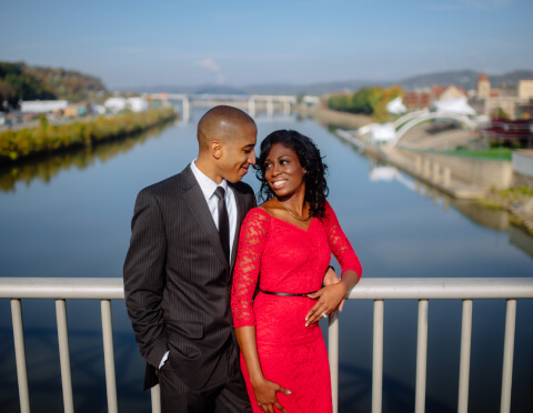 An elegantly dressed couple stands on a bridge with a picturesque river and cityscape behind them, sharing a loving gaze, highlighting their formal attire against the urban landscape.