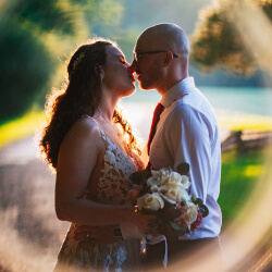 A couple shares a tender kiss in sunlight that filters through a green backdrop.