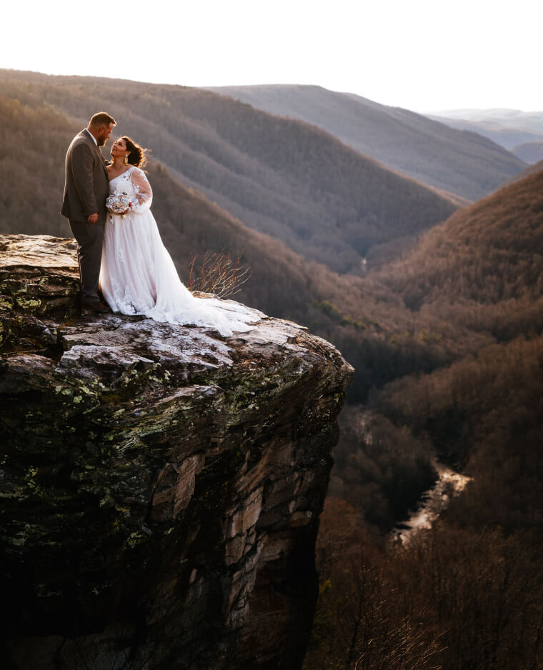 A bride and groom stand on a rocky cliff, overlooking a forested valley at sunset.