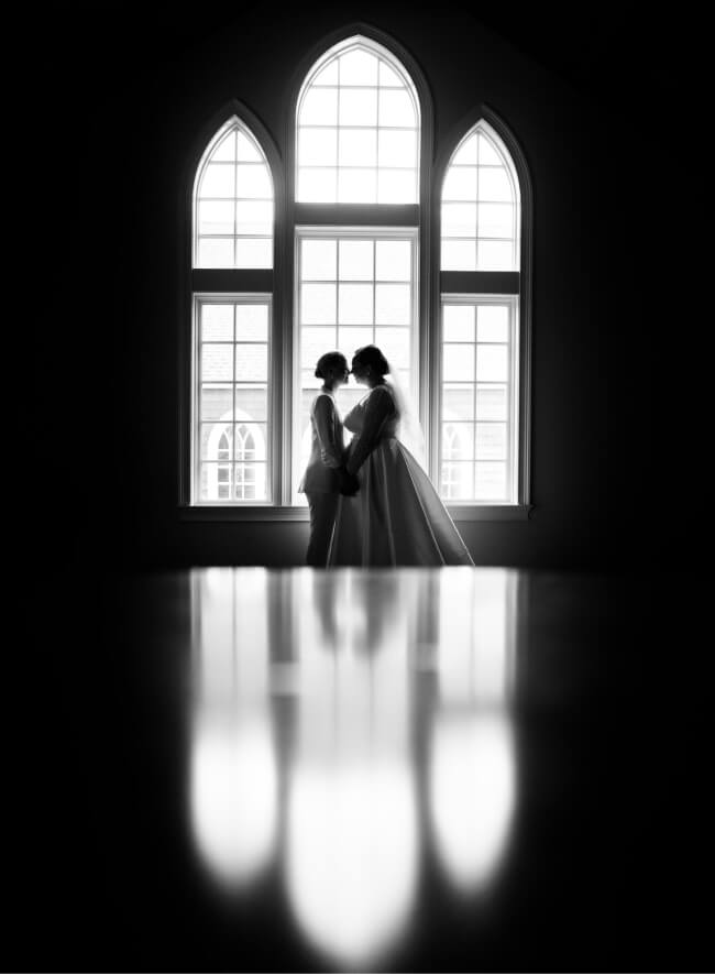 The silhouetted profiles of two brides stand before large arched windows.