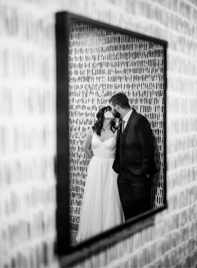 A bride and groom share a kiss, framed in a geometrically patterned mirror that with a textured brick wall.