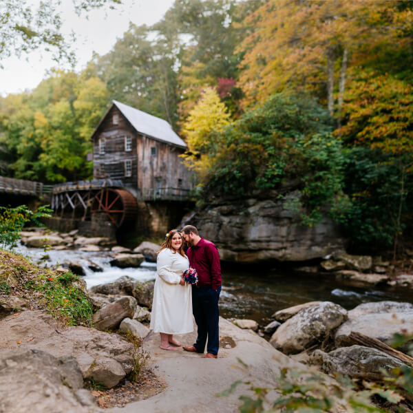 A couple stands together in front of a historic mill during the fall season.