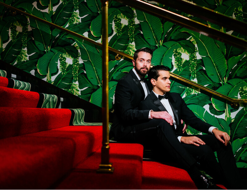 Two grooms in black-tie attire seated on a red staircase, with a bold, tropical leaf pattern in the background.