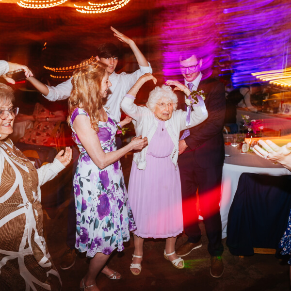 An elderly woman dances happily, surrounded by family and friends with bright lights adding motion and color to the celebration.