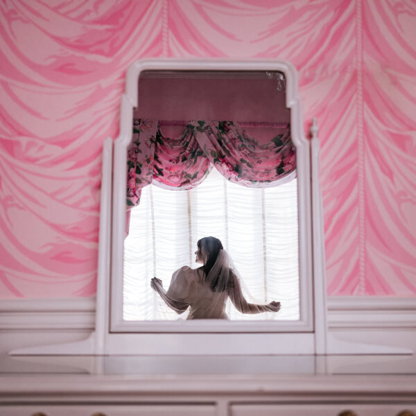 A woman stands in a dress framed by a mirror, contrasting with the pink patterned wallpaper.