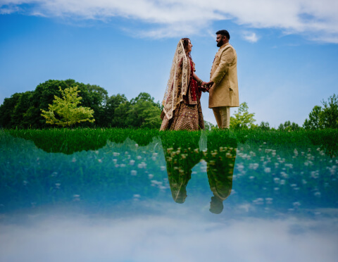 A couple dressed in traditional South Asian wedding attire stands hand in hand, with a perfect reflection in the water beneath them, set against a green landscape with a clear blue sky.