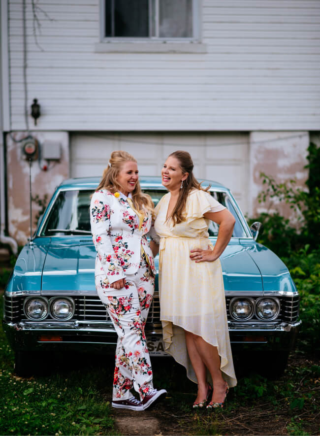 Two brides, one in a floral jumpsuit and the other in a yellow dress, share a laugh together in front of a classic teal car.