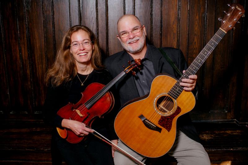 Two people play their instruments smiling in front of a dark wooden wall.