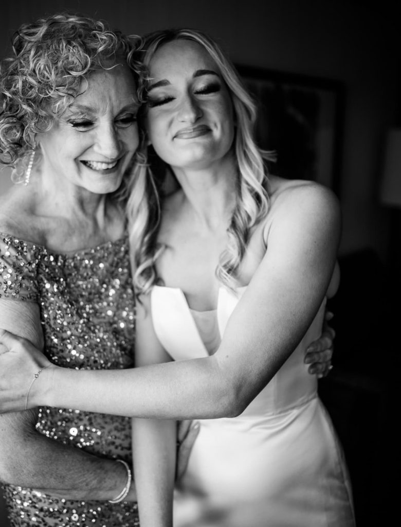 A tender black and white photograph shows a bride in a strapless gown holding her mom in a sparkling dress, both with closed eyes and joyful expressions.