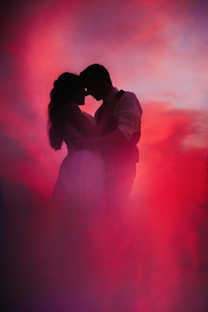 A romantic silhouette of a couple holding one another, set against a dramatic sky with hues of pink and red clouds.