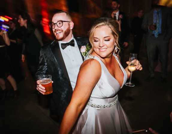 A bride in a white dress with a happy smile dances with a groom in a black velvet tuxedo, enjoying a fun moment at their wedding reception.