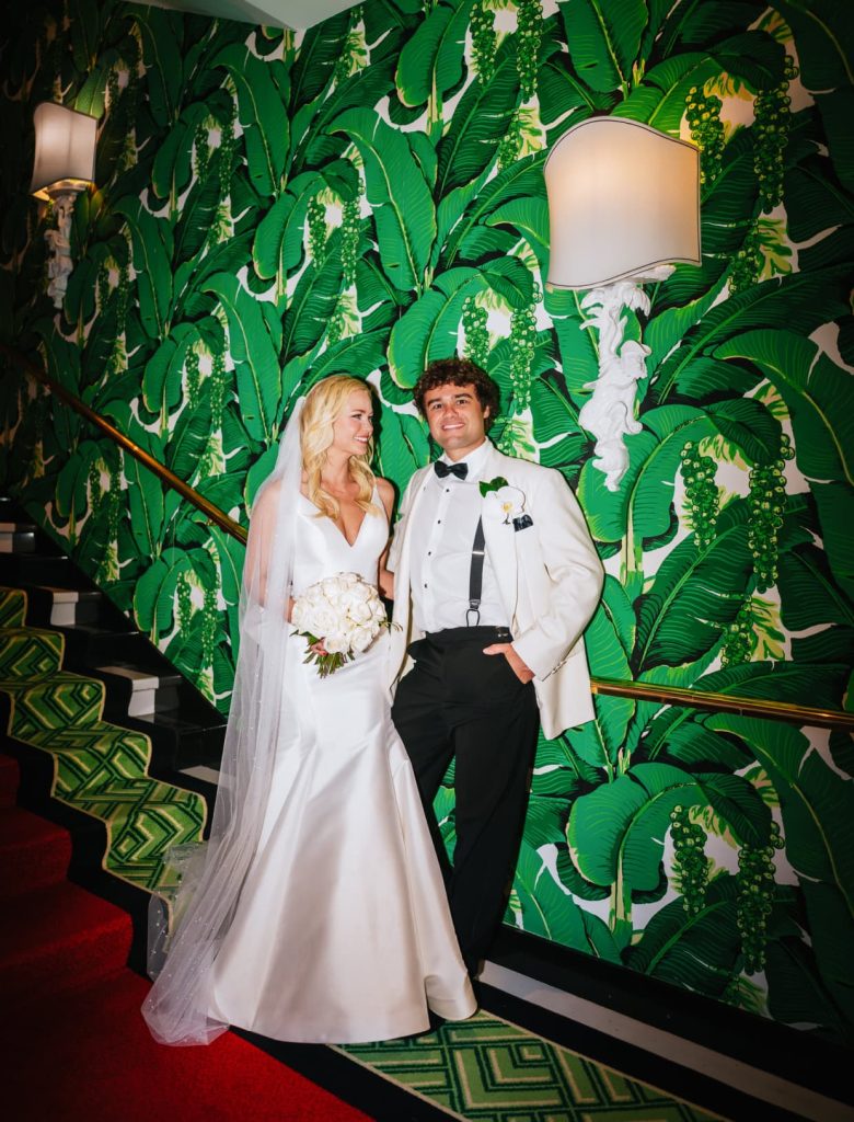 A wedding couple stands on an unique staircase, the bride in white and the groom in a white jacket, against a bold green tropical leaf-patterned wallpaper.