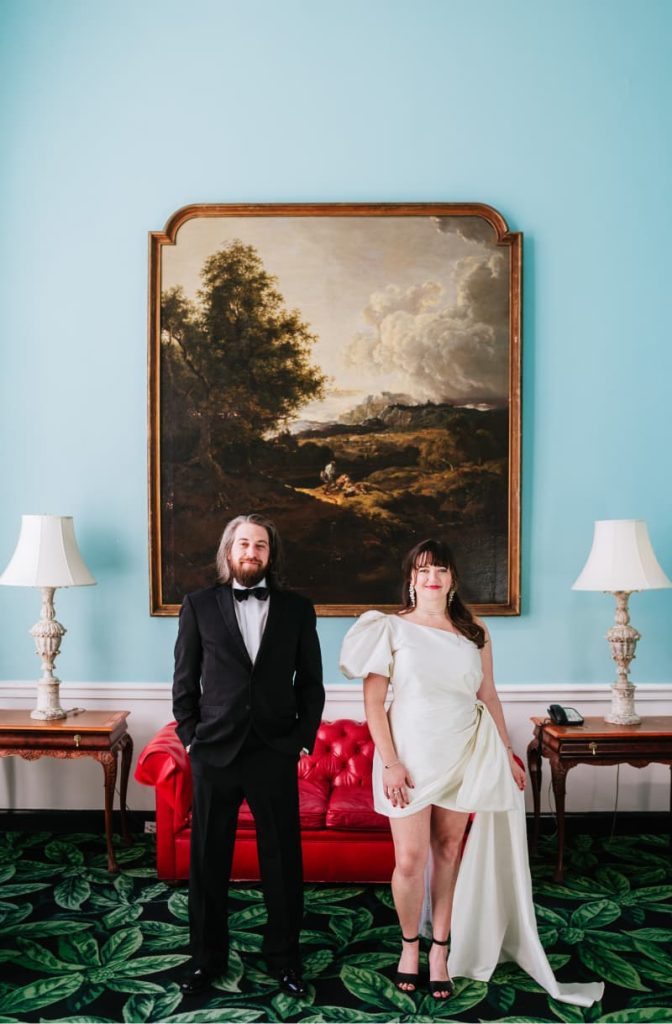 A man in a black tuxedo and a woman in an asymmetrical white dress stand before an elegant, gold-framed landscape painting, in a room with teal walls and red furniture.