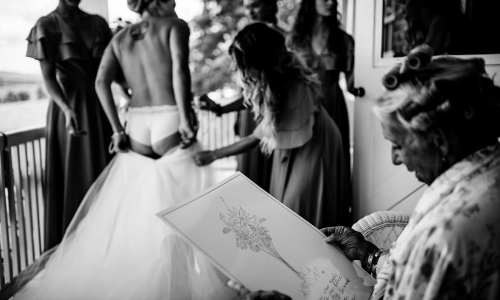 In a black and white photo, a bride gets help with her dress from bridesmaids, while an elderly woman reads a card in the foreground.