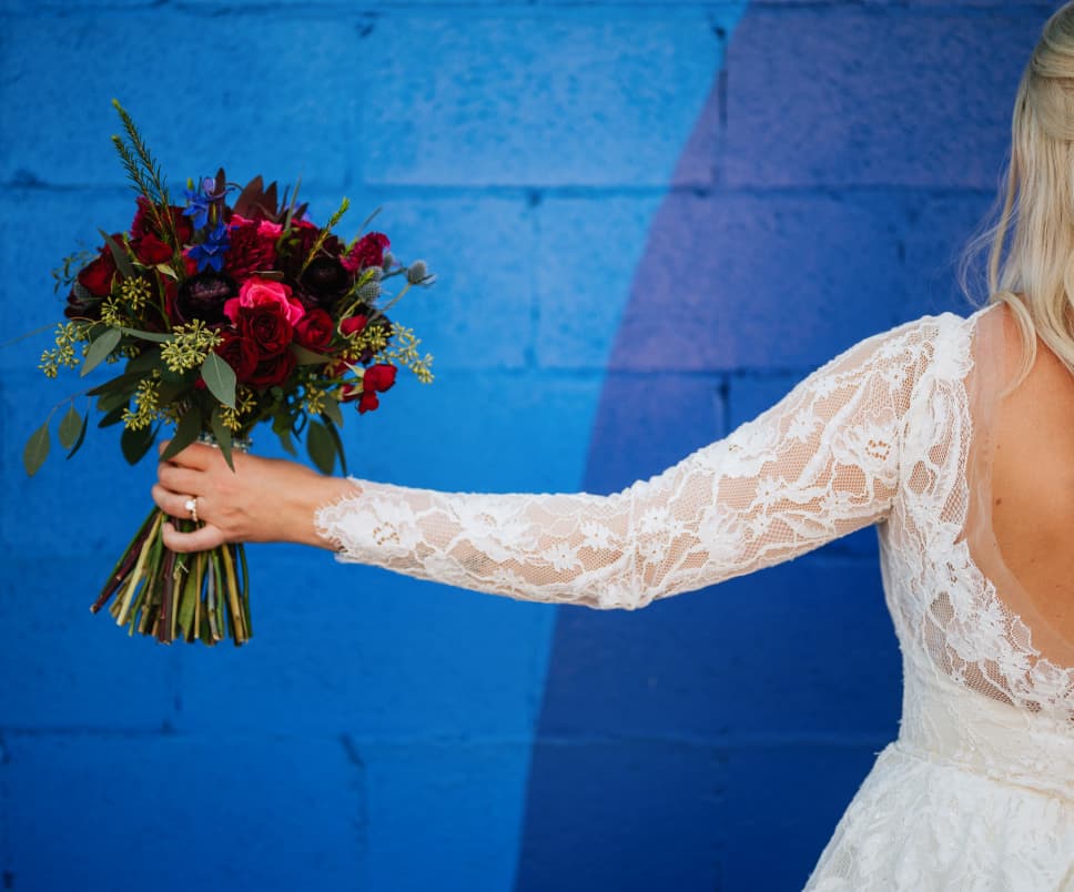 A bride in a lacy wedding dress extends her arm, showing a bouquet of deep red roses against a cobalt blue wall backdrop.