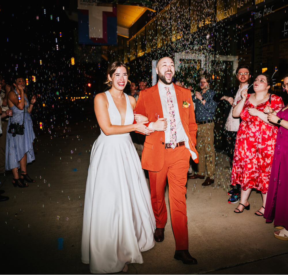 A happy bride in a white dress and a groom in a bright orange suit laugh with a sea of bubbles with wedding guests celebrating around them on a street at night.