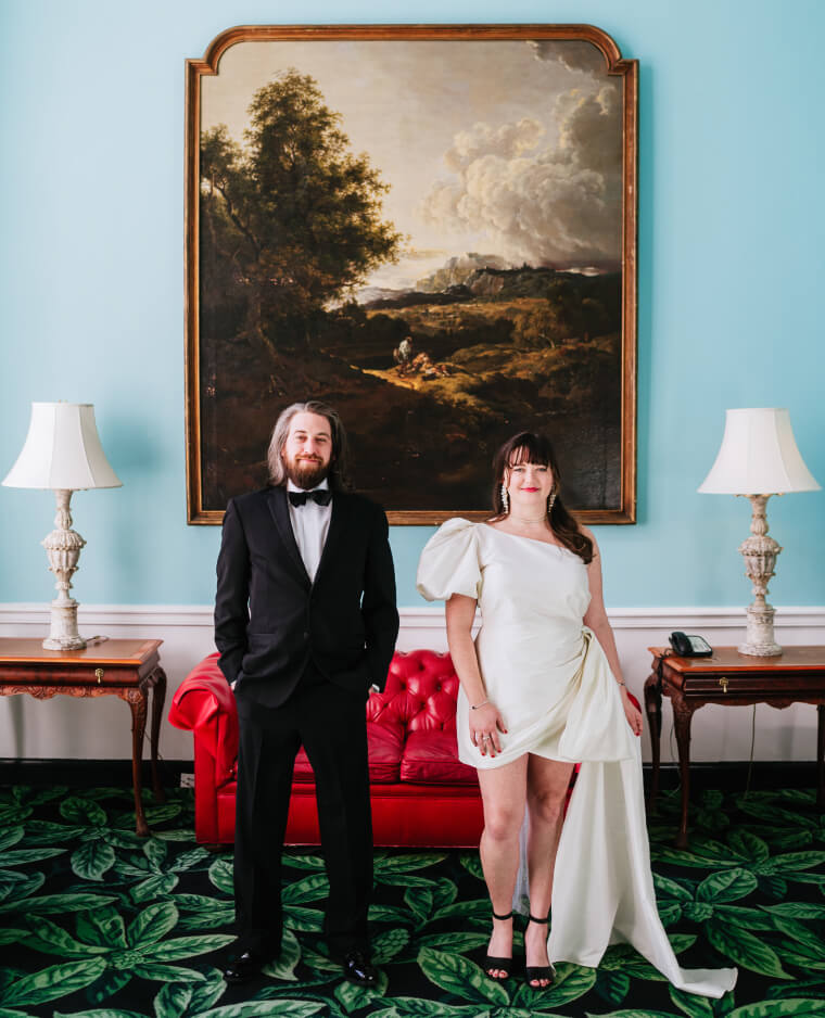 In an elegantly decorated room with blue walls and a classic painting, a couple stands confidently, one person in a tuxedo and the other in a chic dress.