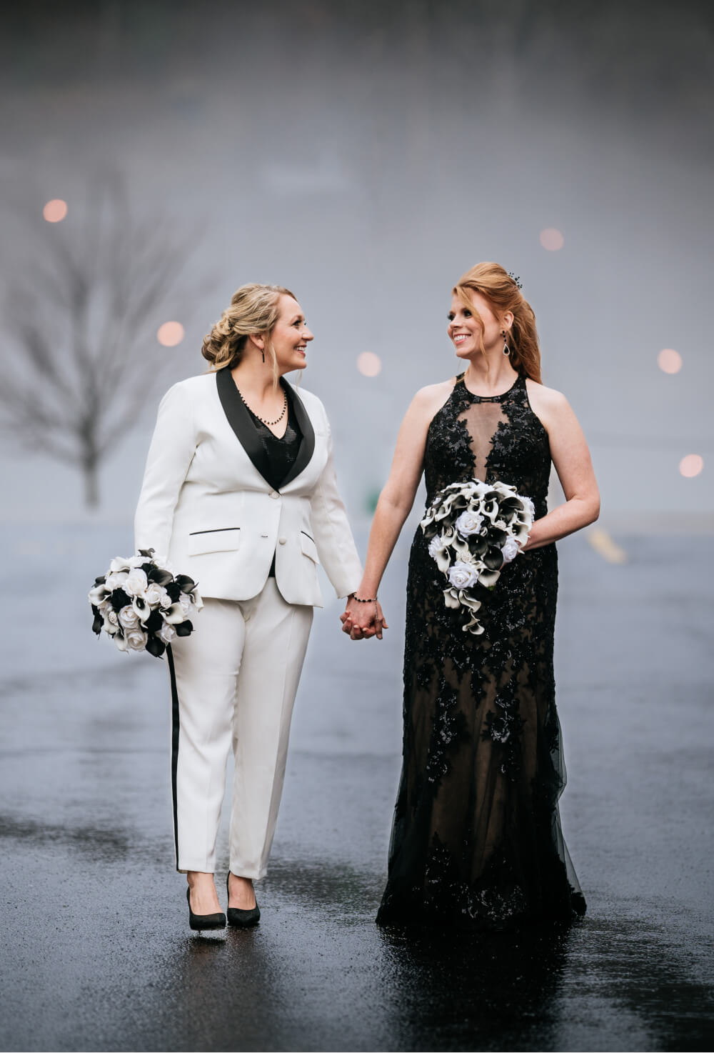 Two brides, one in a white suit and the other in an elegant black gown, holding hands and smiling at each other with a soft, misty background.