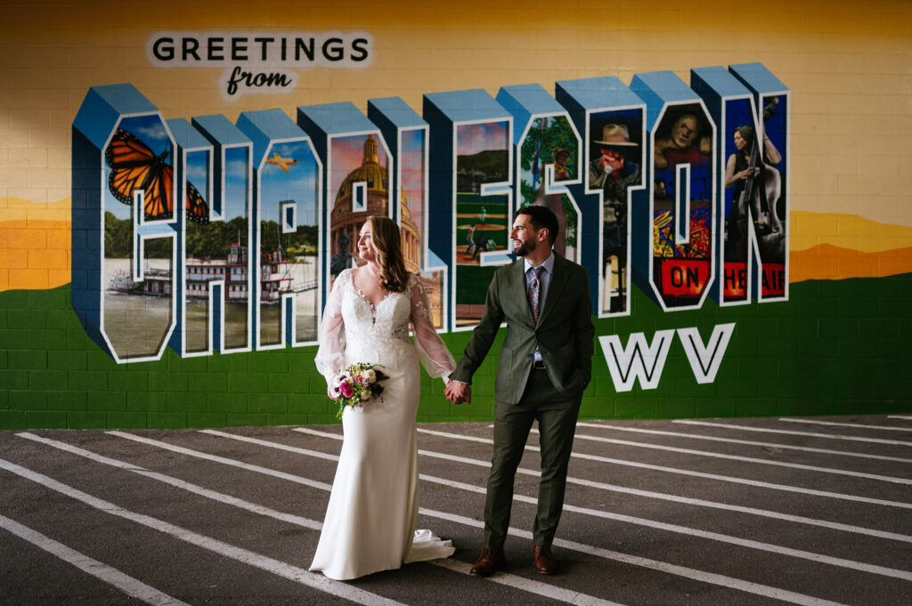 A smiling bride and groom hold hands in front of a vibrant Charleston, WV mural.