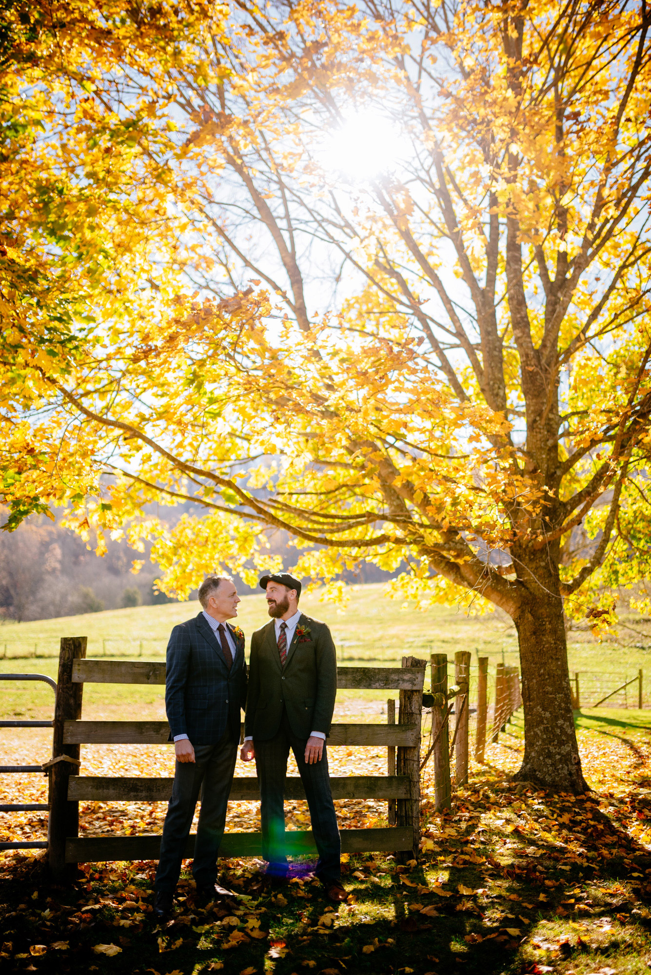 A couple in coordinated autumnal attire stands by a wooden fence under a golden yellow tree.