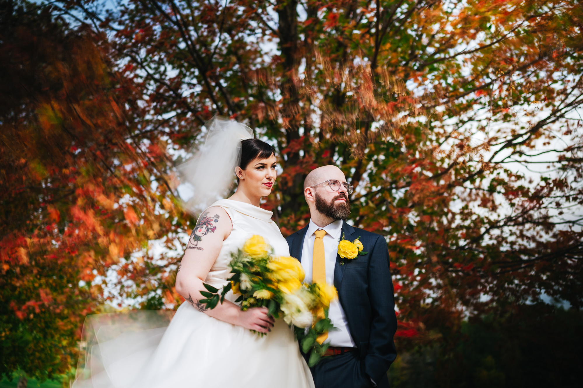 A bride and groom, with the bride showcasing unique tattoos, stand before autumn trees.