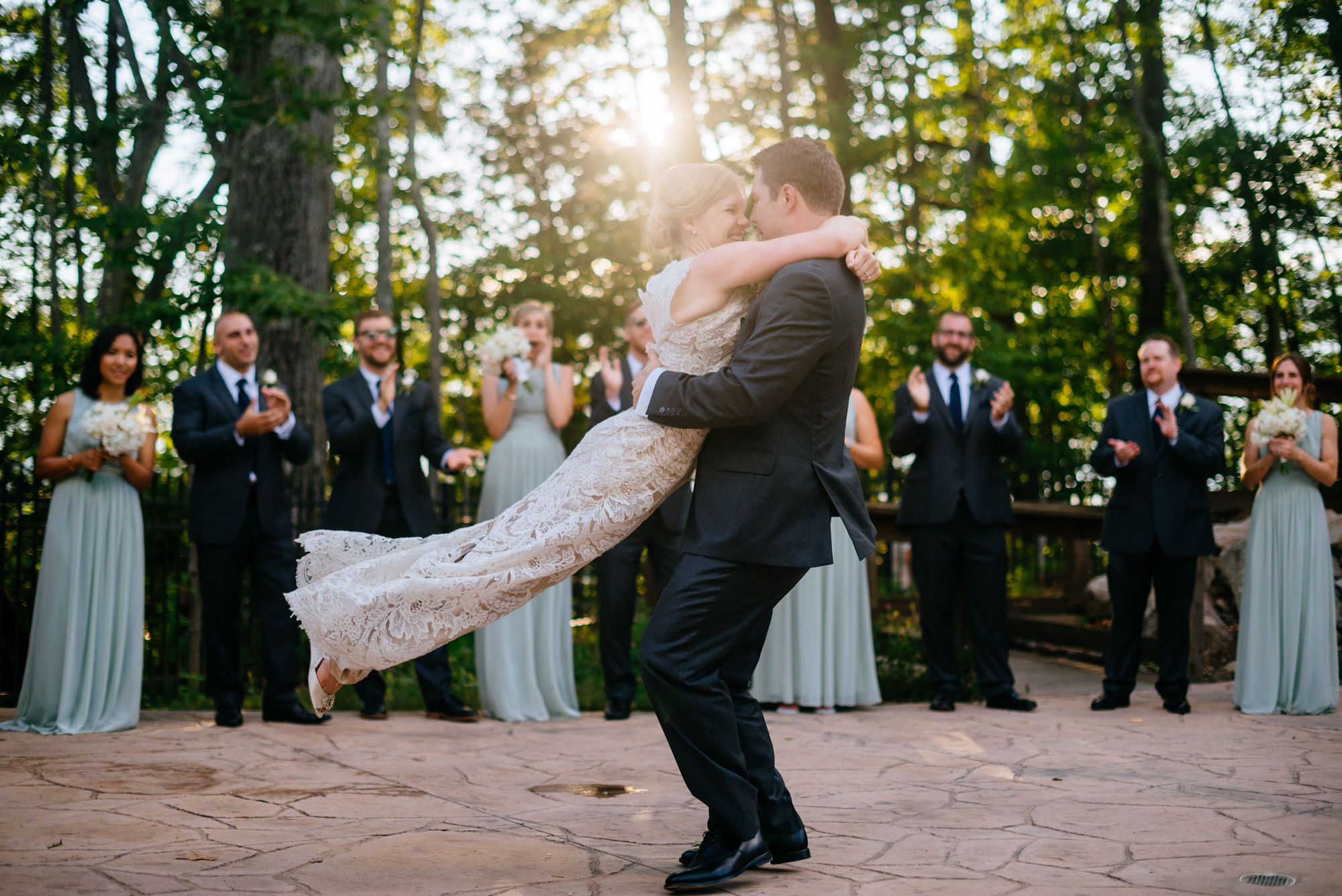 A bride being twirled by her groom on a dance floor outdoors, surrounded by applauding bridesmaids and groomsmen.