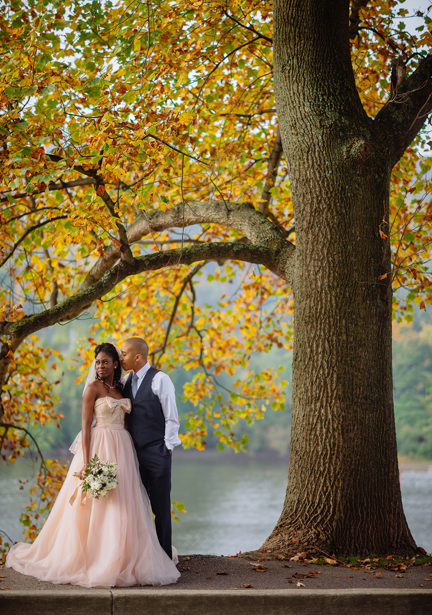 A newlywed couple posing under autumm trees, with the bride in a peach dress and the groom in a vest and tie, by a river.