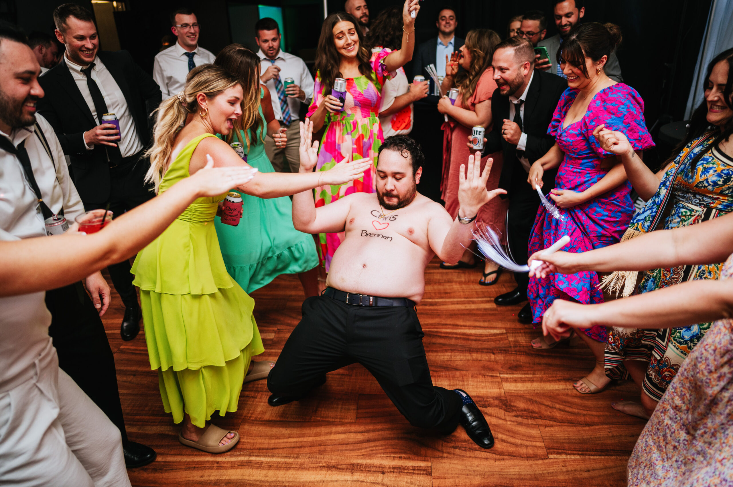 A shirtless man dancing at a wedding reception with guests' hands raised around him, the name 'Chris Berman' written on his chest.