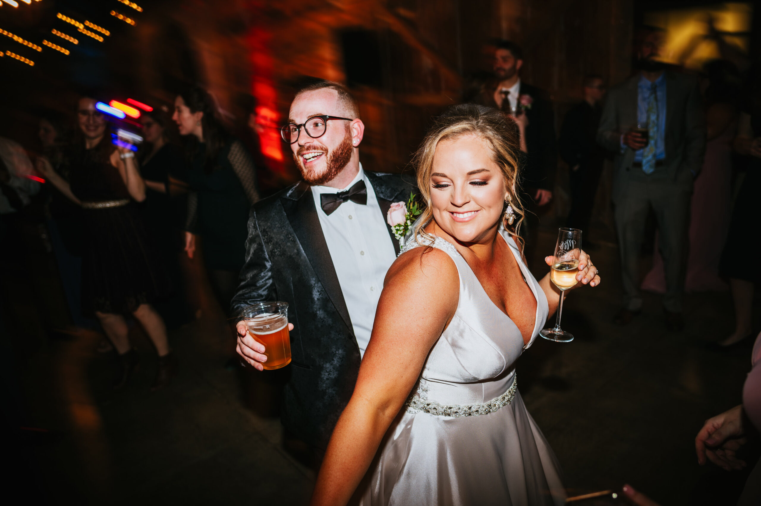 A smiling couple dancing at a wedding reception, with the man holding a beer and the woman holding a glass of wine.