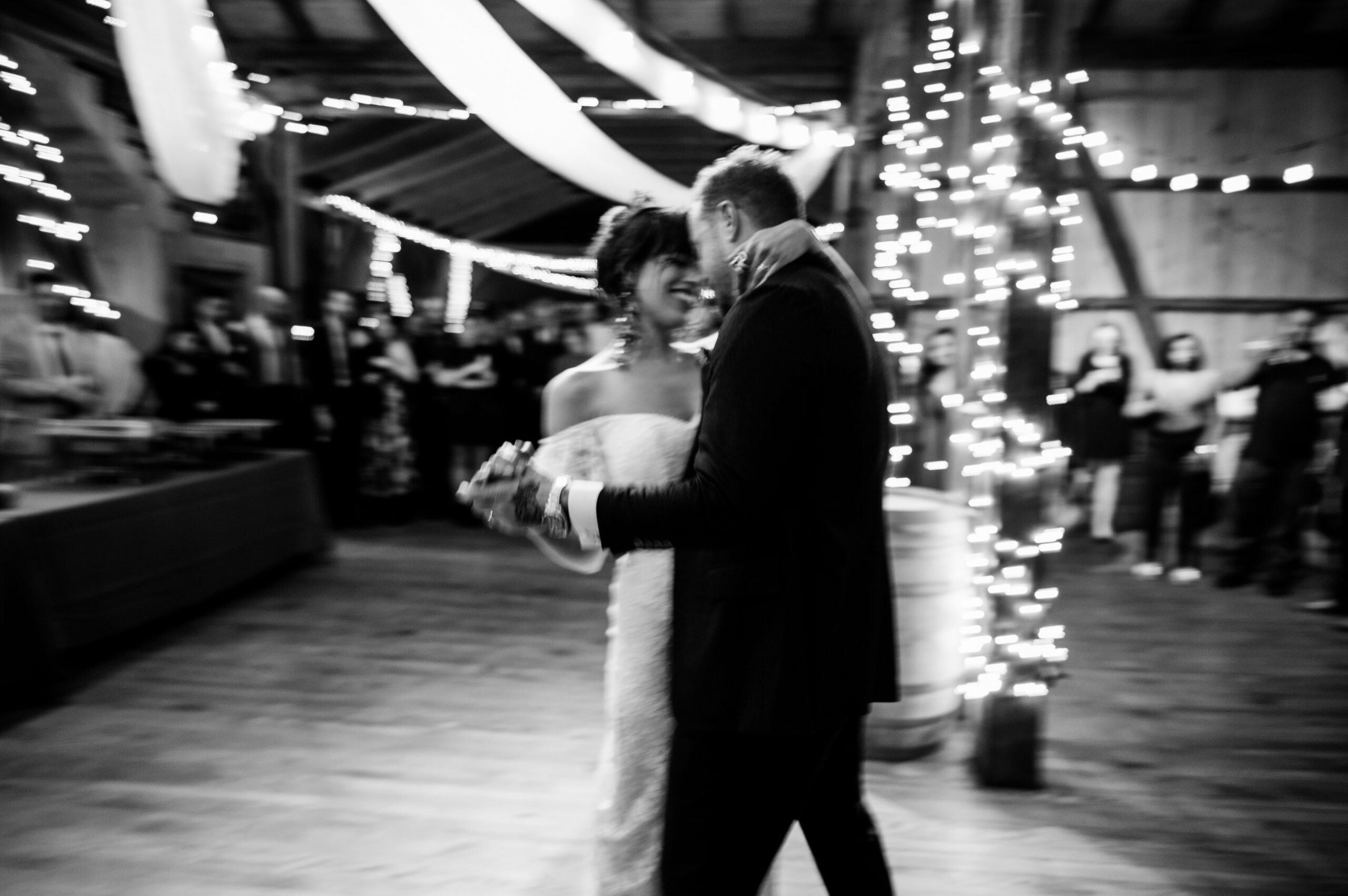 A bride and groom in blurred motion, sharing a dance in a warmly lit barn with guests and string lights in the background.