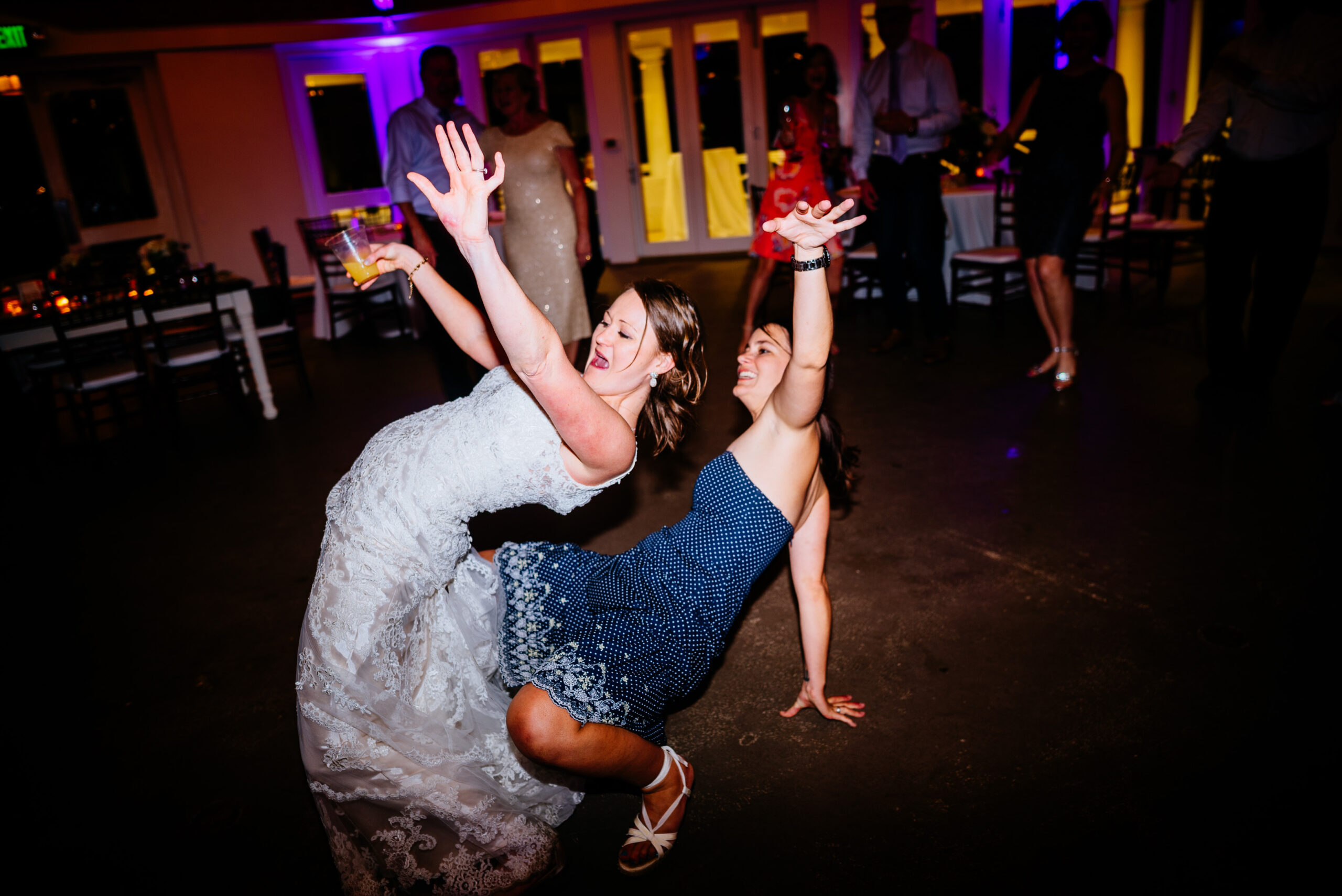 A dance floor moment at a wedding reception, with a bride in a lace gown and a guest in a navy blue dress, dancing back-to-back.