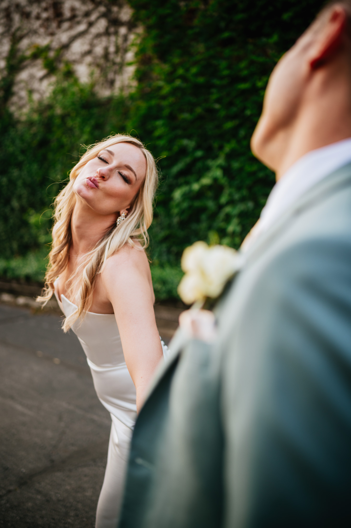 A bride puckering for a kiss towards the viewer with her eyes playfully closed, a blurred groom in the foreground holding a boutonniere.