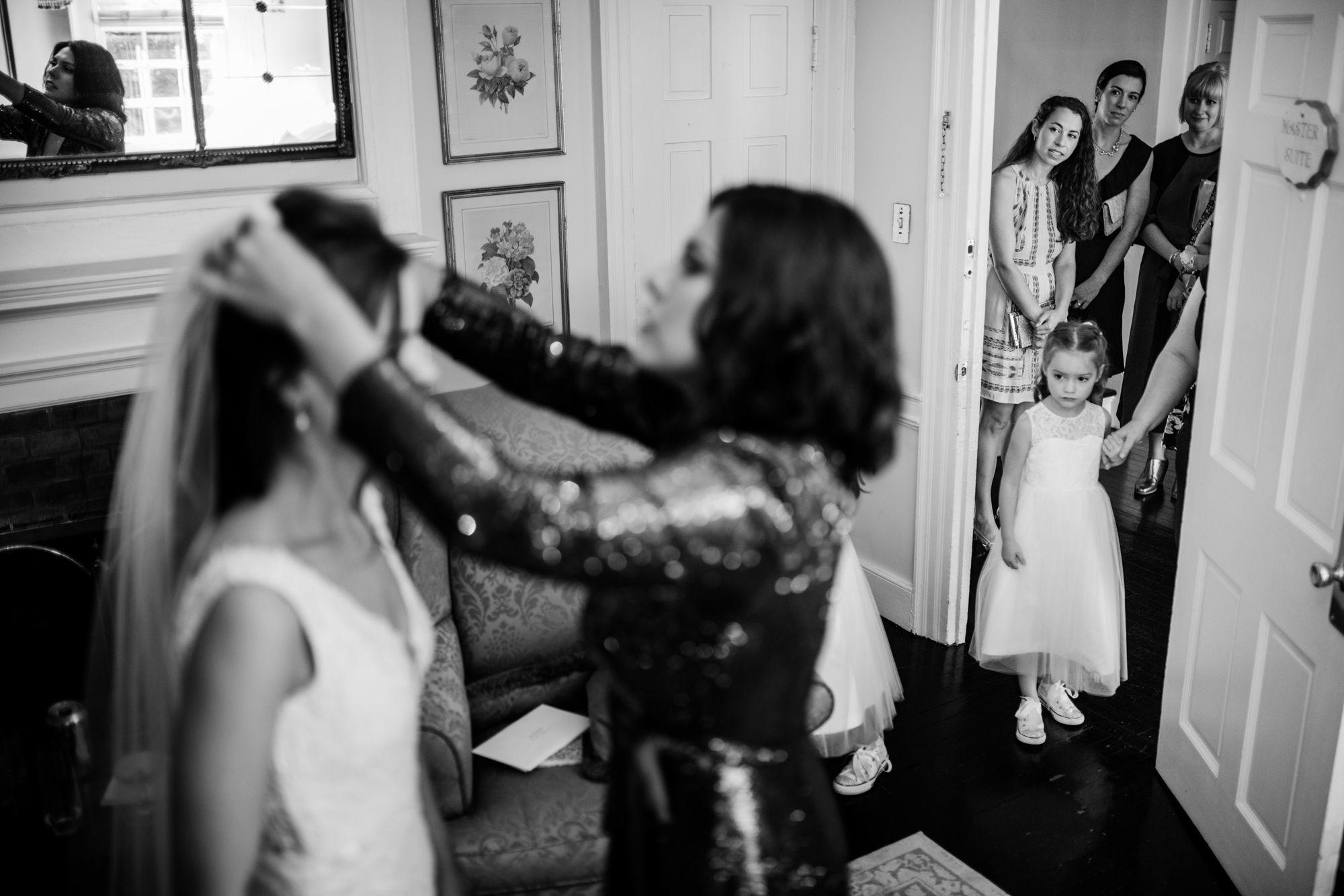 A behind-the-scenes black-and-white image of a bride getting ready, with a flower girl looking on and guests in the doorway.