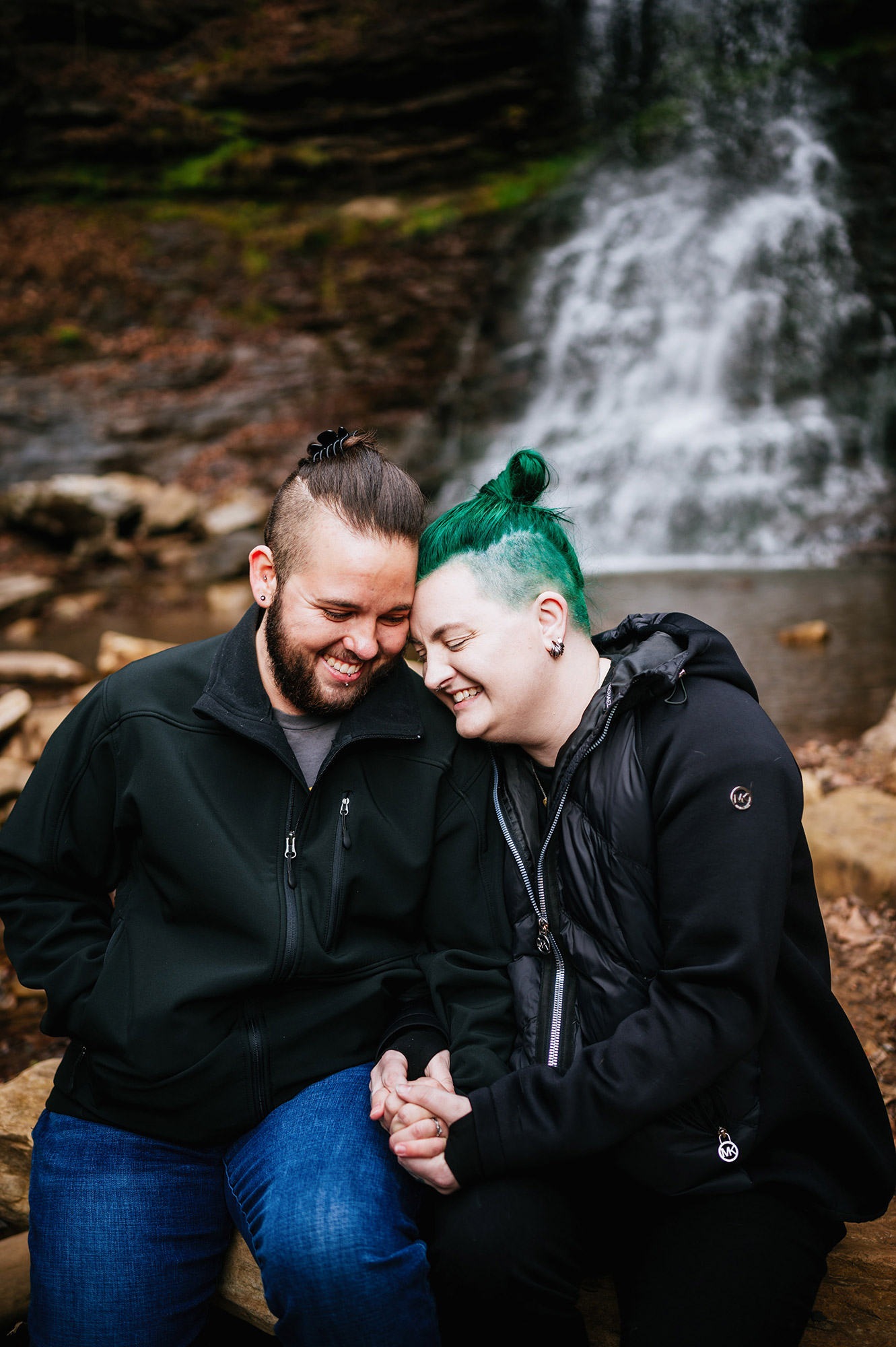 A couple sits closely together, holding hands and smiling by a waterfall.
