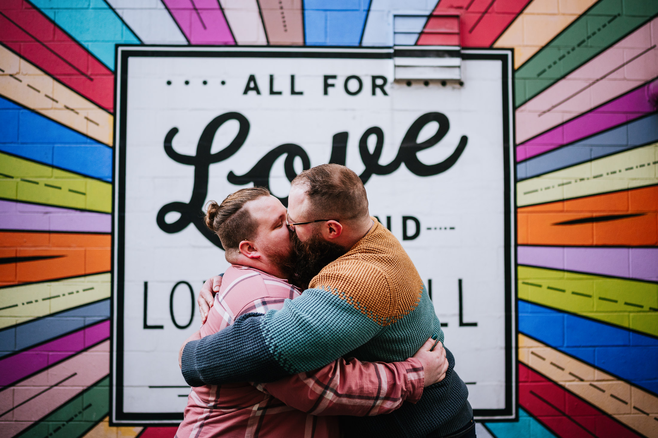 Two partners embracing in front of a colorful mural stating "All For Love And Love For All".